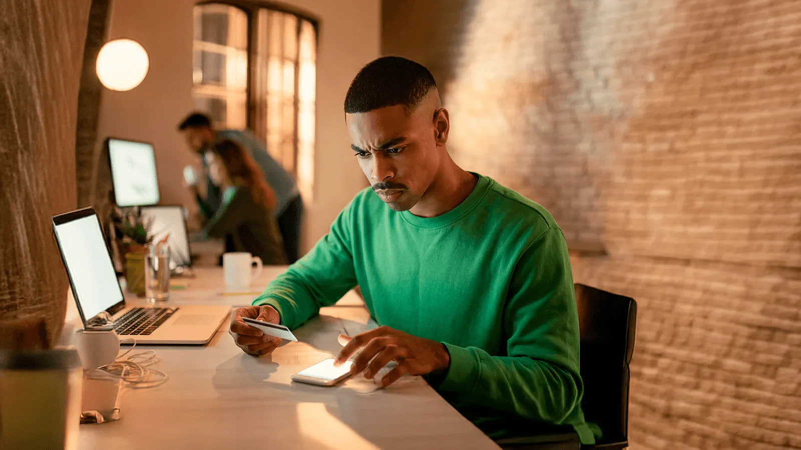 Homem realizando uma compra online pelo celular e cartão de crédito com expressão de frustração durante o processo de checkout em um escritório moderno.